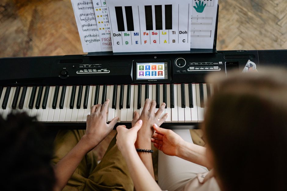 A woman guides a student's hands on a keyboard piano, emphasizing musical instruction.