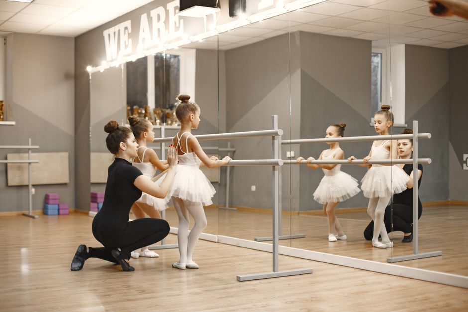 Ballet instructor helps young girls in tutus practice at a mirrored barre.
